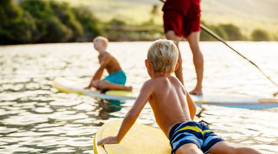 A child laying on the front of a paddle board, looking towards two other people on a paddle board.